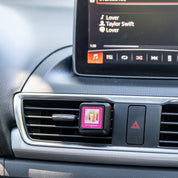 A Bridgewater Candle Company Lets Celebrate Auto Vent Clip is attached to the car dashboard, offering a floral fragrance as Lover by Taylor Swift plays on the infotainment screen.