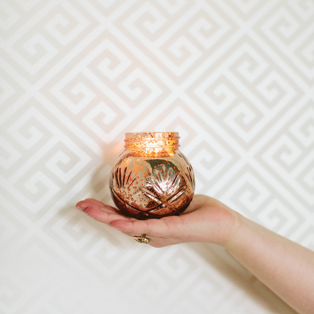 A hand with a ring holds a small, decorative glass candle holder with a lit candle. The candleholder has a gold and bronze leaf design. The background features a white wall with a geometric pattern.