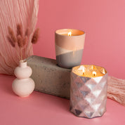 Two lit Bridgewater Candle Company Sweet Grace Collection Candle #041s in decorative holders are styled on and beside a concrete block next to a small white vase with dried pink flowers, all against a pink background with soft draped fabric.