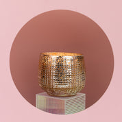 A textured, gold-colored round vase rests on a translucent pedestal against a pink backdrop, echoing the elegance of Bridgewater Candle Companys Sweet Grace Collection Candle #051—our signature best-seller.
