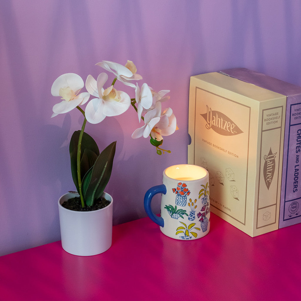 A white potted orchid, a colorful mug with a Bridgewater Candle Company Sweet Grace Collection Candle #054 inside, and a Yahtzee game box sit on a pink surface against a lavender wall.