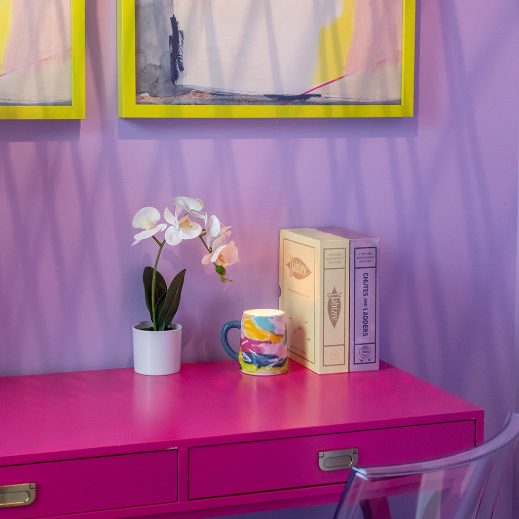 A pink desk with a white potted orchid, colorful mug, Bridgewater Candle Companys Sweet Grace Collection Candle #055, and two books sits against a purple wall. Yellow-framed abstract art hangs above as window shadows fall across the scene.