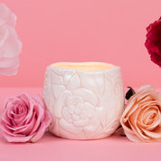 The Sweet Grace Collection Candle #057 by Bridgewater Candle Company features a white, floral-patterned ceramic holder with soy-blend wax, surrounded by roses on a pink backdrop for a luxurious home fragrance accent.