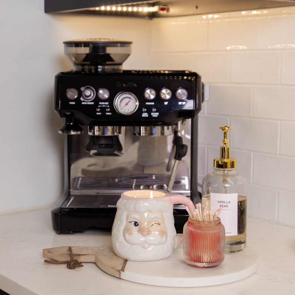 A coffee machine sits on a kitchen counter next to a round tray holding a Bridgewater Candle Company Sweet Grace Santa Mug, a pink jar with matches, and a vanilla bean soap with gold pump. White subway tile backsplash adds holiday charm.