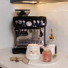 A coffee machine sits on a kitchen counter next to a round tray holding a Bridgewater Candle Company Sweet Grace Santa Mug, a pink jar with matches, and a vanilla bean soap with gold pump. White subway tile backsplash adds holiday charm.