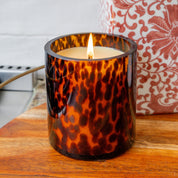 A lit Bridgewater Candle Company Sweet Grace Collection Candle #064 in a brown tortoise glass holder sits on a wooden surface beside a decorative red and white floral container.