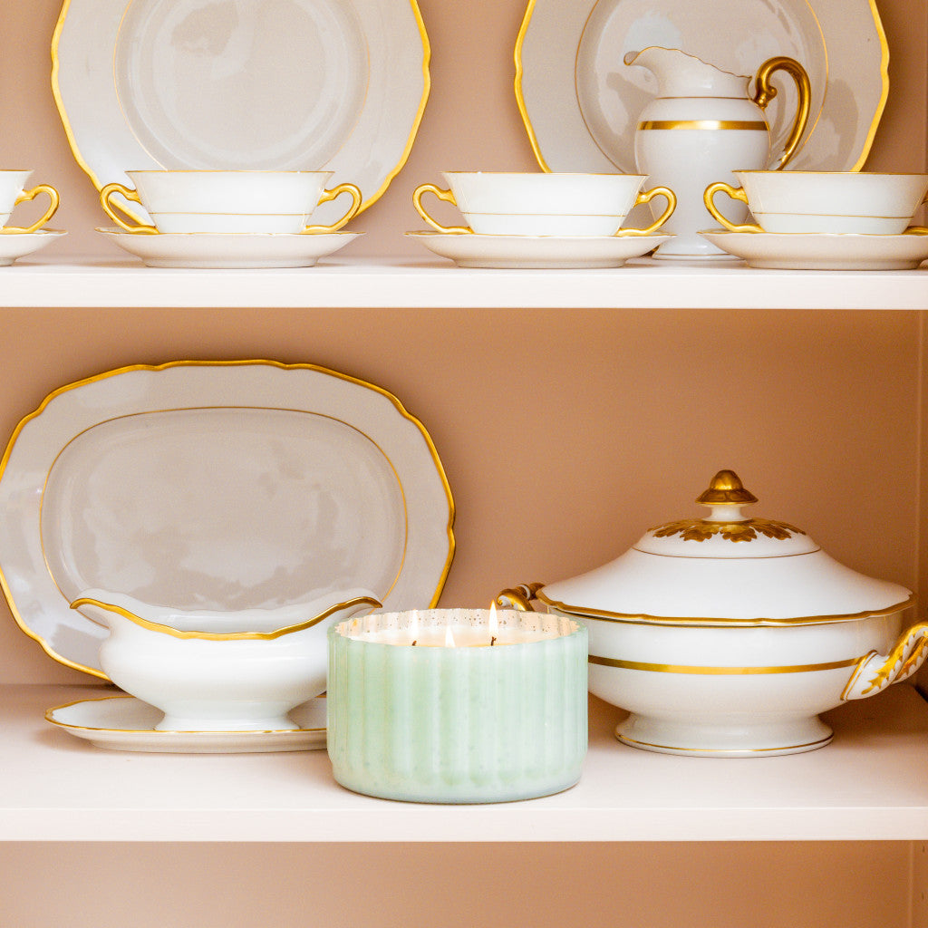 A shelf displaying elegant white porcelain dishes with gold trim—cups, saucers, plates, gravy boat, lidded tureen, oval platter—and a Bridgewater Candle Company Sweet Grace Collection Candle #065 in an antique green ribbed vessel.