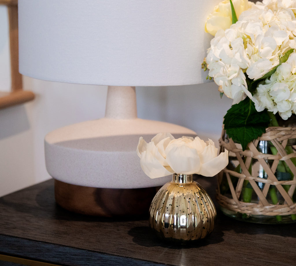 A table features a modern lamp, a gold vase with a white flower, a basket of white hydrangeas, and the Bridgewater Candle Company Sweet Grace Mercury Flower Diffuser adding an elegant home fragrance.