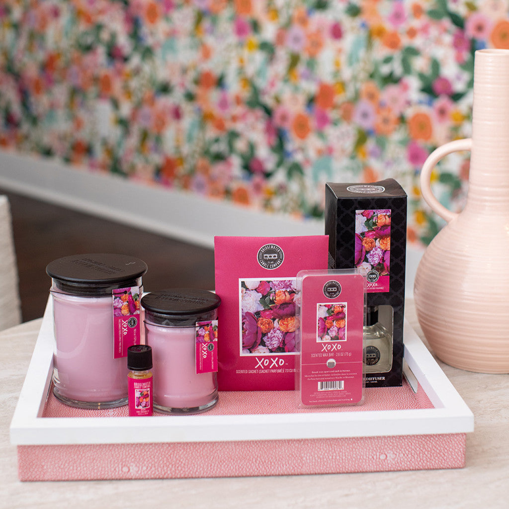 A white tray holds pink jar candles, a small essential oil bottle, wax melts, and a boxed Bridgewater Candle Company XOXO Reed Diffuser on a table with a peach vase and floral wallpaper in the background.