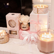 A cozy scene features lit candles, pink and white ribboned gift boxes, a Bridgewater Candle Company Sweet Grace Mercury Jar Candle, and a boxed flower diffuser by the fireplace, all glowing with warm soy-blend wax ambiance.