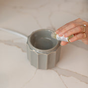 A hand pours home fragrance oils into a gray scalloped mug on a marble countertop beside the Bridgewater Candle Company Wax Warmer-Grey Banded. The person is wearing gold rings.