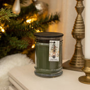 A Bridgewater Candle Company Festive Frasier 18oz Large Jar Candle with a black lid rests on a white table by a brass candlestick before a lit, ornamented Christmas tree, filling the room with its festive scent.
