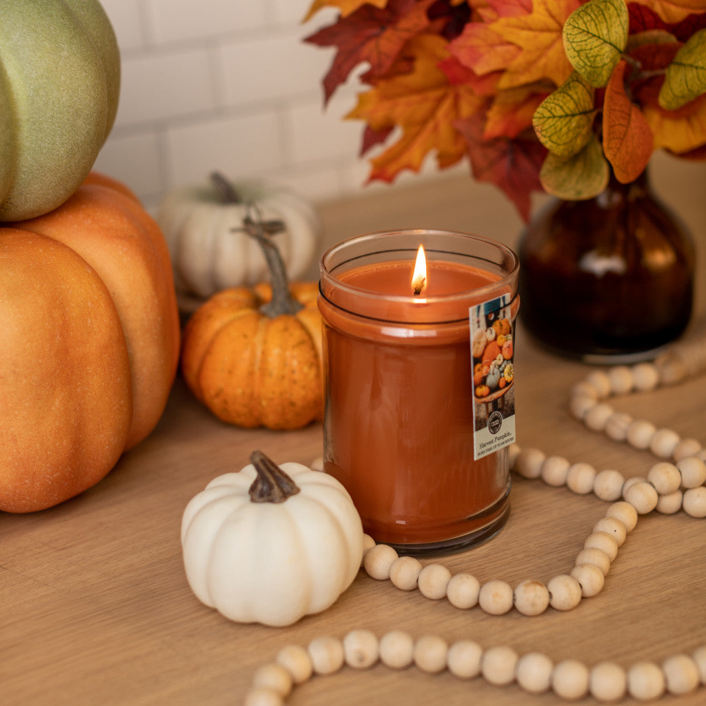 The Bridgewater Candle Company Harvest Pumpkin 8oz Jar Candle sits in a glass jar on a wooden surface with small pumpkins, wooden beads, and autumn leaves, creating a cozy fall atmosphere.