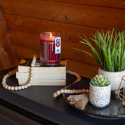 A lit Welcome Home 18oz Large Jar Candle by Bridgewater Candle Company sits on stacked books beside wooden beads, while potted succulents and a plant rest nearby on a dark tabletop in a cozy, wooden room infused with its fragrance.