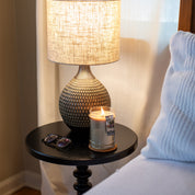 A Bridgewater Candle Company Afternoon Retreat 18oz Large Jar Candle, eyeglasses, and a decorative lamp sit on a round black nightstand next to a bed with white bedding, near a softly lit window with sheer curtains.
