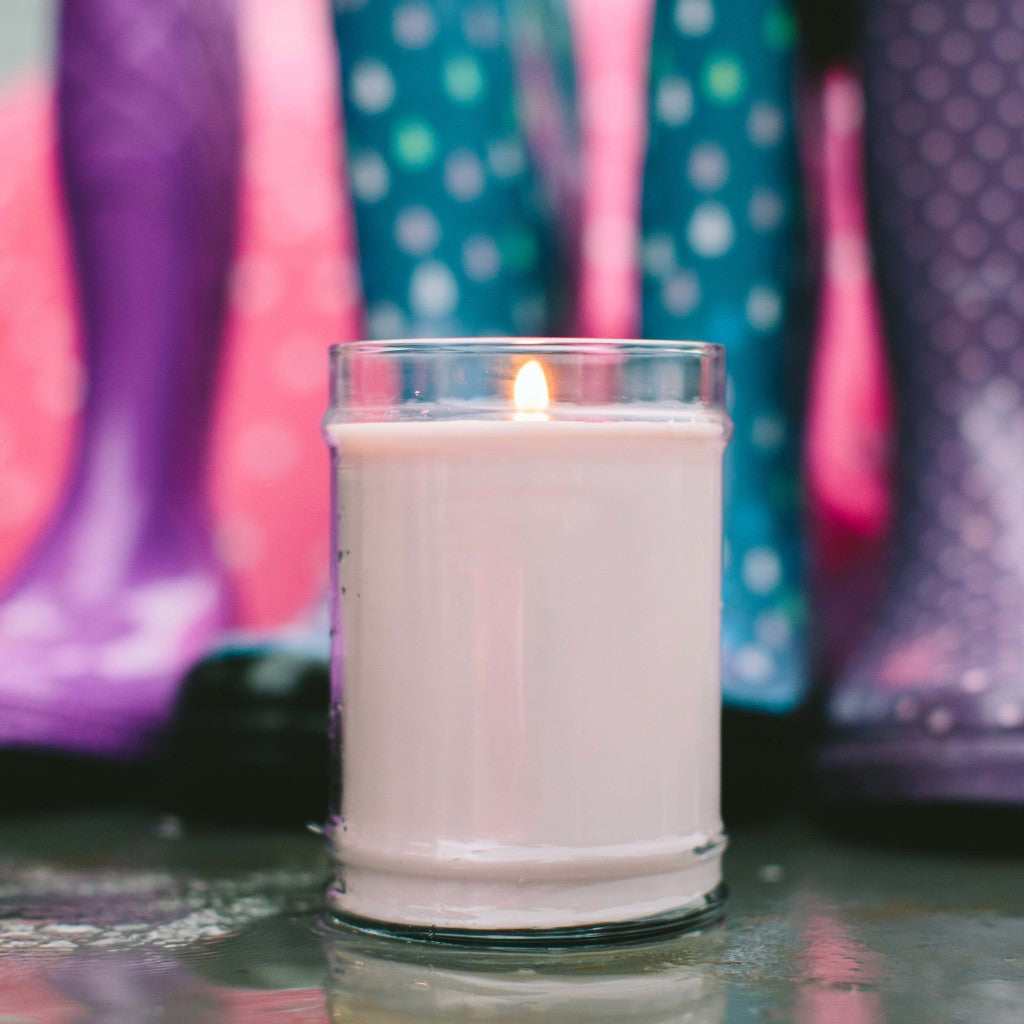 A lit Bridgewater Candle Company Kiss in the Rain 18oz Large Jar Candle sits on a wet surface, with blurred colorful polka dot rain boots in the background, capturing the fragrances fresh ambiance.