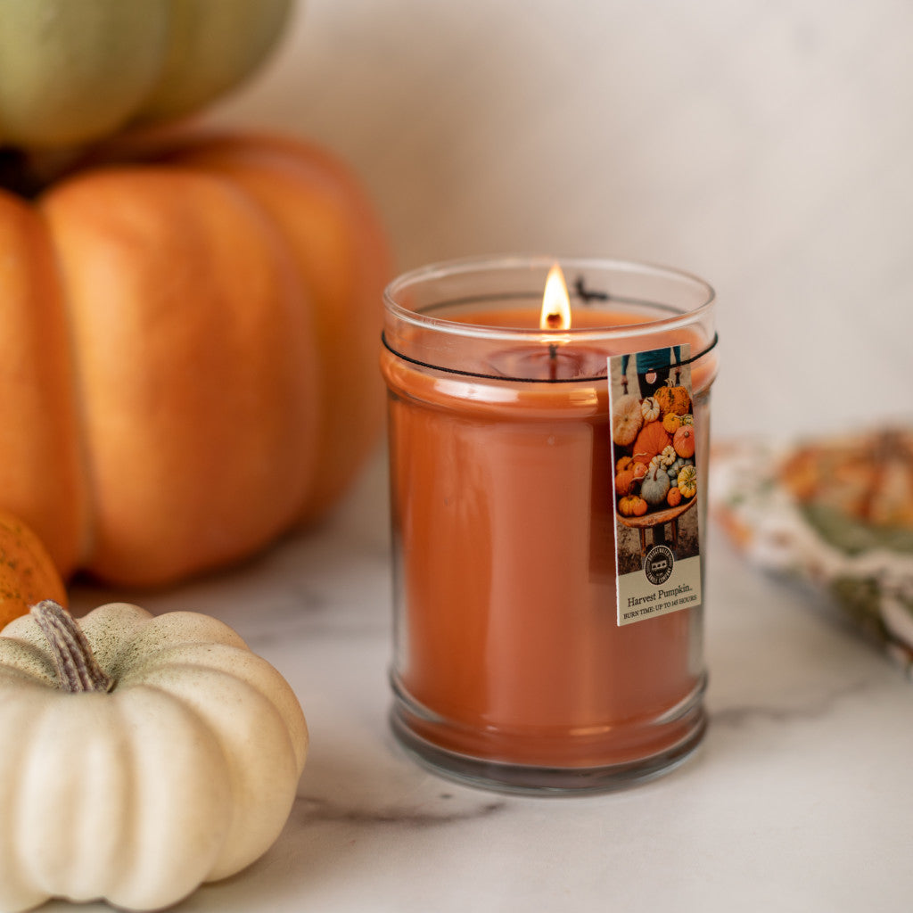 A Bridgewater Candle Company Harvest Pumpkin 18oz Large Jar Candle sits on a marble surface, surrounded by small white and orange pumpkins with a patterned fabric nearby.