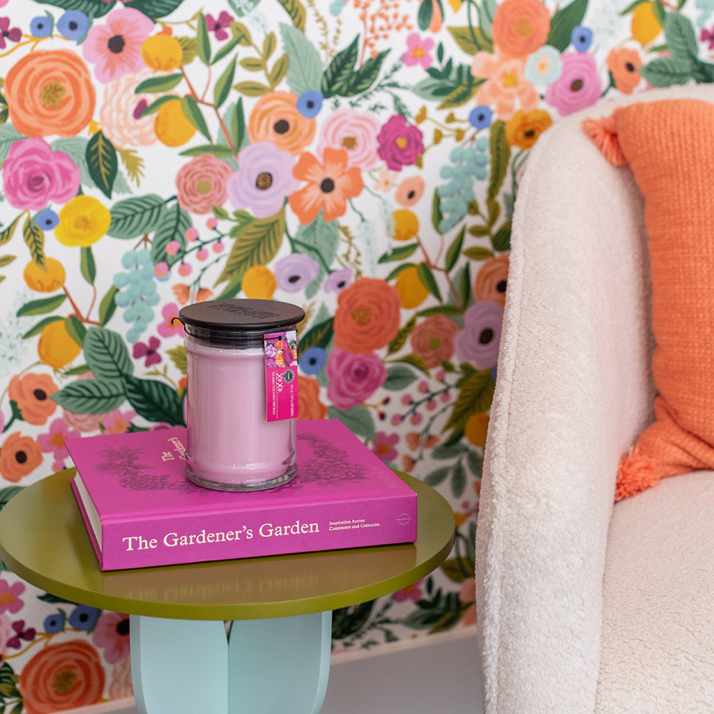 A pink Bridgewater Candle Company XOXO 18oz Large Jar Candle and the pink book The Gardener’s Garden rest on a green side table by a white chair with an orange pillow, set against vibrant floral wallpaper.