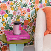A pink Bridgewater Candle Company XOXO 18oz Large Jar Candle and the pink book The Gardener’s Garden rest on a green side table by a white chair with an orange pillow, set against vibrant floral wallpaper.