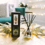 The Bridgewater Candle Company Festive Frasier Reed Diffuser, with fiber reeds in a clear glass bottle and a decorative box, is displayed on a gold-patterned table among pine branches and fairy lights; a green chair is blurred in the background.