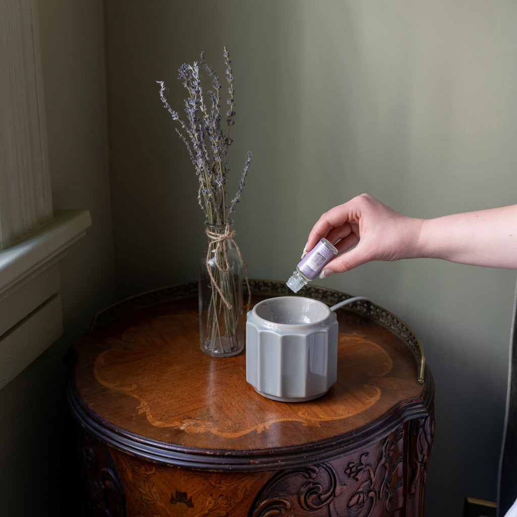 A hand pours essential oil into a white ceramic diffuser on a wooden table, next to a glass vase with dried lavender.