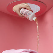 A hand pours Bridgewater Candle Company Sweet Grace 6oz Laundry Detergent through a round pink opening onto a pink cloth, with a soft pink background for fabric-safe cleaning.