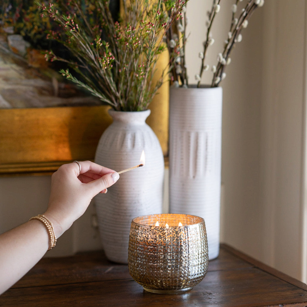 A hand lights a Sweet Grace Collection Candle #051 by Bridgewater Candle Company, made with soy-blend wax, on a wooden table, with two white vases filled with branches in the textured glass background.
