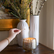 A hand lights a Sweet Grace Collection Candle #051 by Bridgewater Candle Company, made with soy-blend wax, on a wooden table, with two white vases filled with branches in the textured glass background.