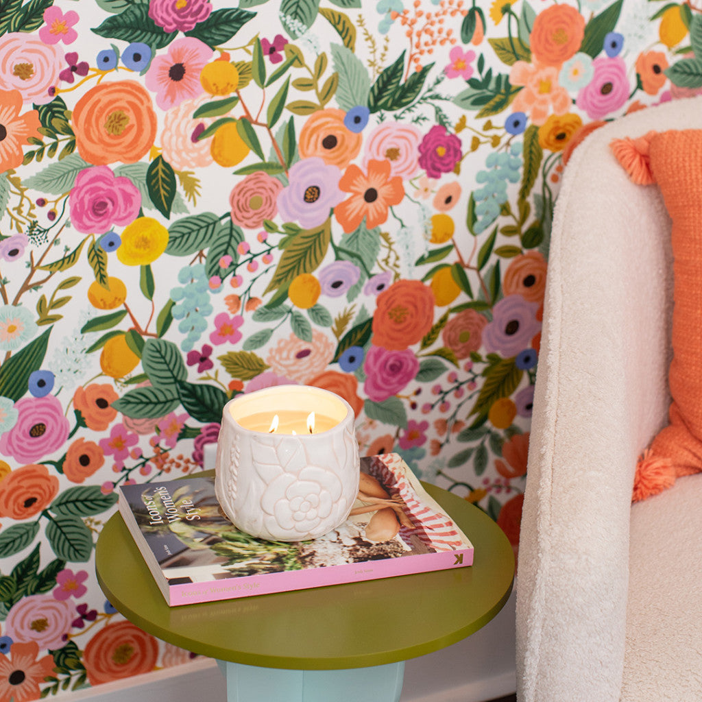 A Bridgewater Candle Company Sweet Grace Collection Candle #057, soy-blend wax, rests atop two magazines on a green side table beside a cream armchair with an orange pillow, framed by vibrant floral wallpaper for a luxurious home fragrance touch.