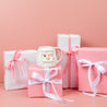 The Bridgewater Candle Company Sweet Grace Santa Mug Candle sits on a pink gift box amid pink and white ribbon-wrapped boxes. Once the candle burns out, reuse the festive Santa mug for cheerful holiday drinks.