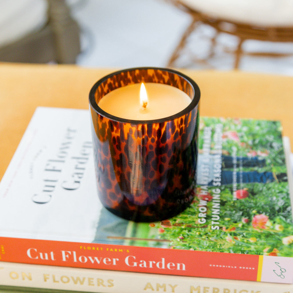 A Sweet Grace Collection Candle #064 by Bridgewater Candle Company, in a brown tortoise glass holder, sits on two stacked Cut Flower Garden books. A blurred sofa and wicker chair are visible in the background.