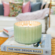 A Bridgewater Candle Company Sweet Grace Collection Candle #065, in an antique green ribbed vessel with three wicks, burns cozily atop The New Design Rules hardcover on a woven table, with a sofa and colorful cushions behind. Crafted from soy-blend wax.