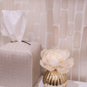 A woven tissue box with a tissue sticking out stands beside a gold vase and the Bridgewater Candle Company Sweet Grace Mercury Flower Diffuser, filling the room with its fragrance against a beige-patterned wall.