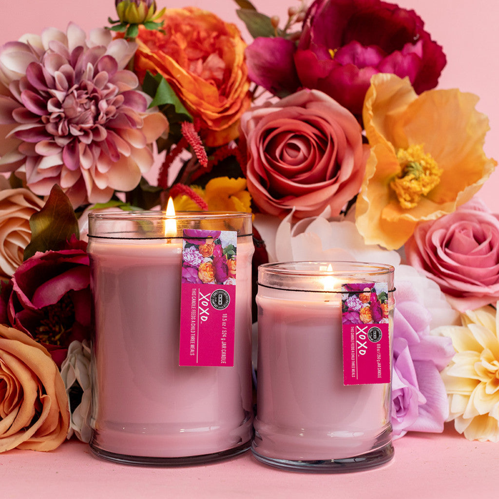 Two Bridgewater Candle Company XOXO 8oz small jar candles with floral-themed labels are lit and placed in front of a colorful arrangement of roses and other flowers on a pink background.