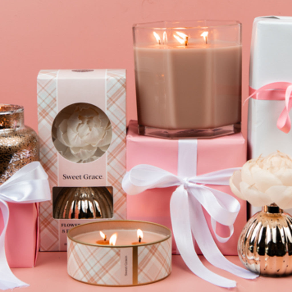 A collection of pink-themed gifts, featuring the Bridgewater Candle Company Sweet Grace Mercury Jar Candle and flower diffusers, some tied with pink and white ribbons, arranged against a soft pink background.