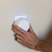 A hand with a gold ring presses the Pura + Bridgewater Sweet Grace Lovers Kit by Bridgewater Candle Company, glowing blue, into a wall outlet—ready to fill the room with Sweet Grace fragrance from this sleek smart diffuser.