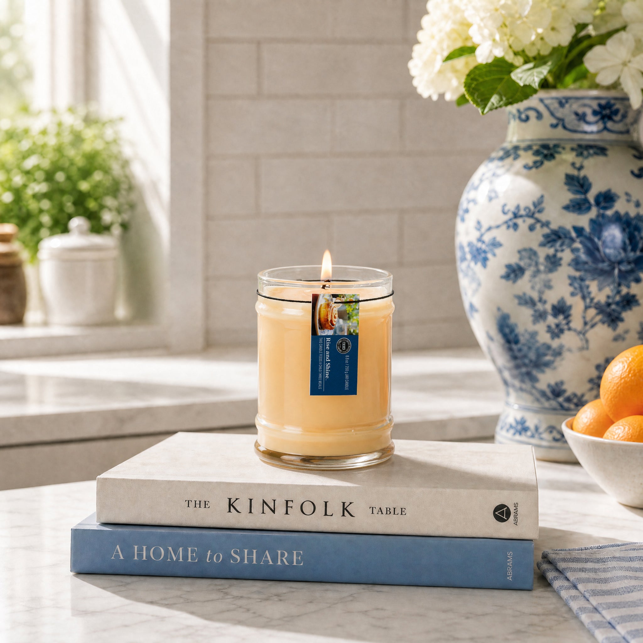 The Rise & Shine 8oz Jar Candle by Bridgewater Candle sits atop two stacked books on a marble countertop. In the background, a blue and white vase with white flowers and a bowl of oranges glow in sunlight streaming through a window.