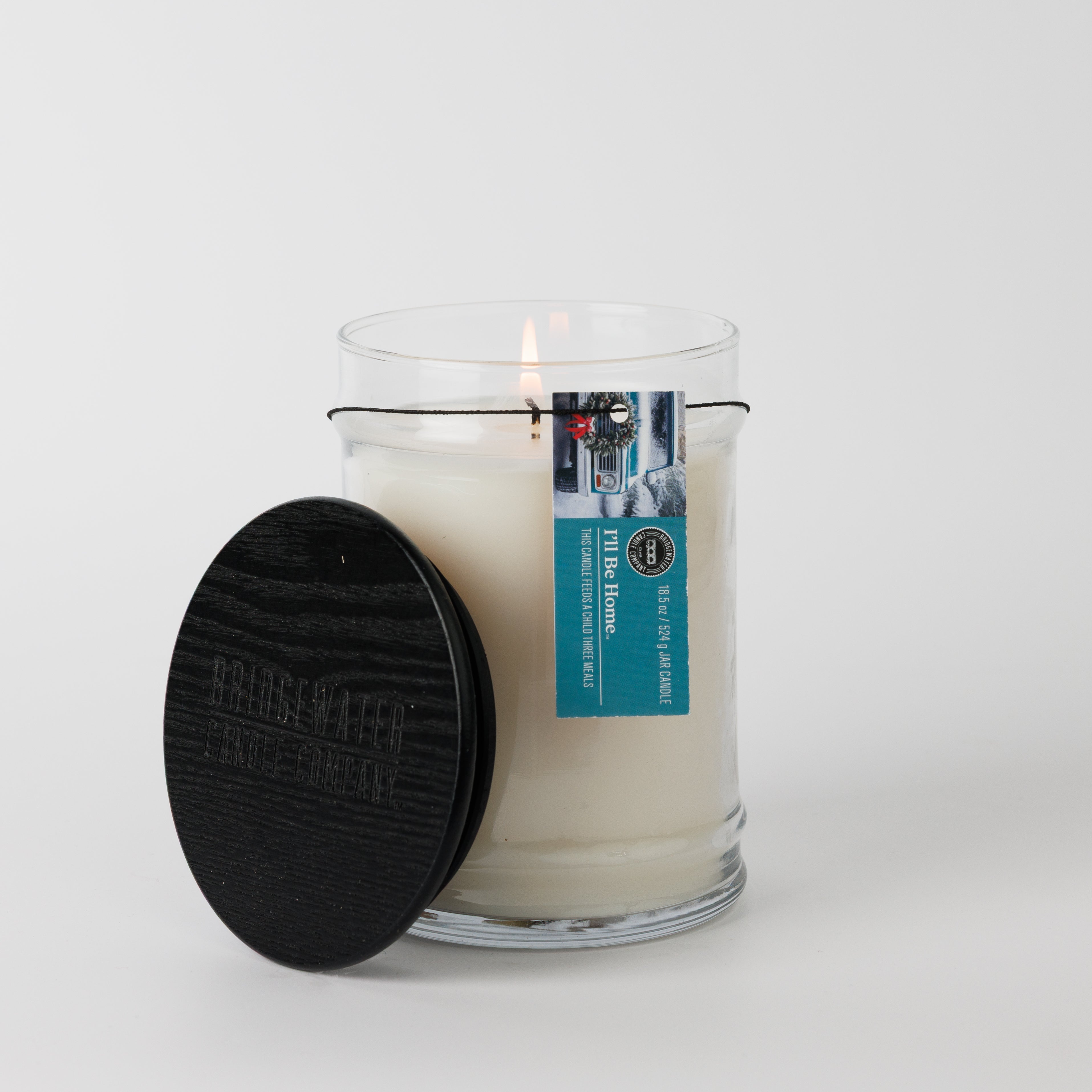 The Bridgewater Candle "I'll Be Home" 18oz Large Jar Candle, made with soy-blend wax, sits in a clear glass jar with a black wooden lid and blue label, filling your room with fragrance against a plain white background.