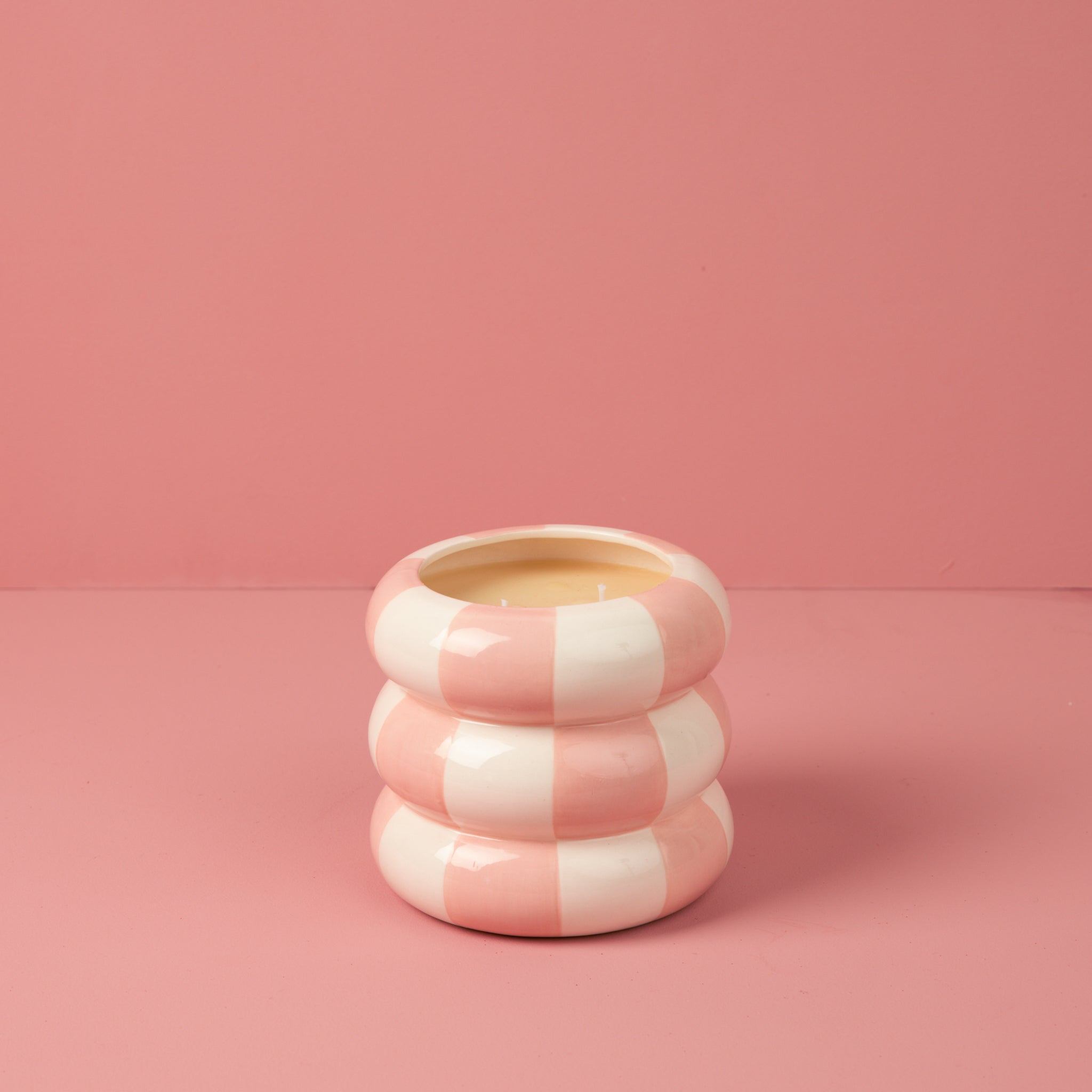 The Sweet Grace Collection Candle #074 by Bridgewater Candle features a pink and white striped, triple-stacked ceramic holder with a cream soy-blend candle inside, shown on a pink surface against a pink background.