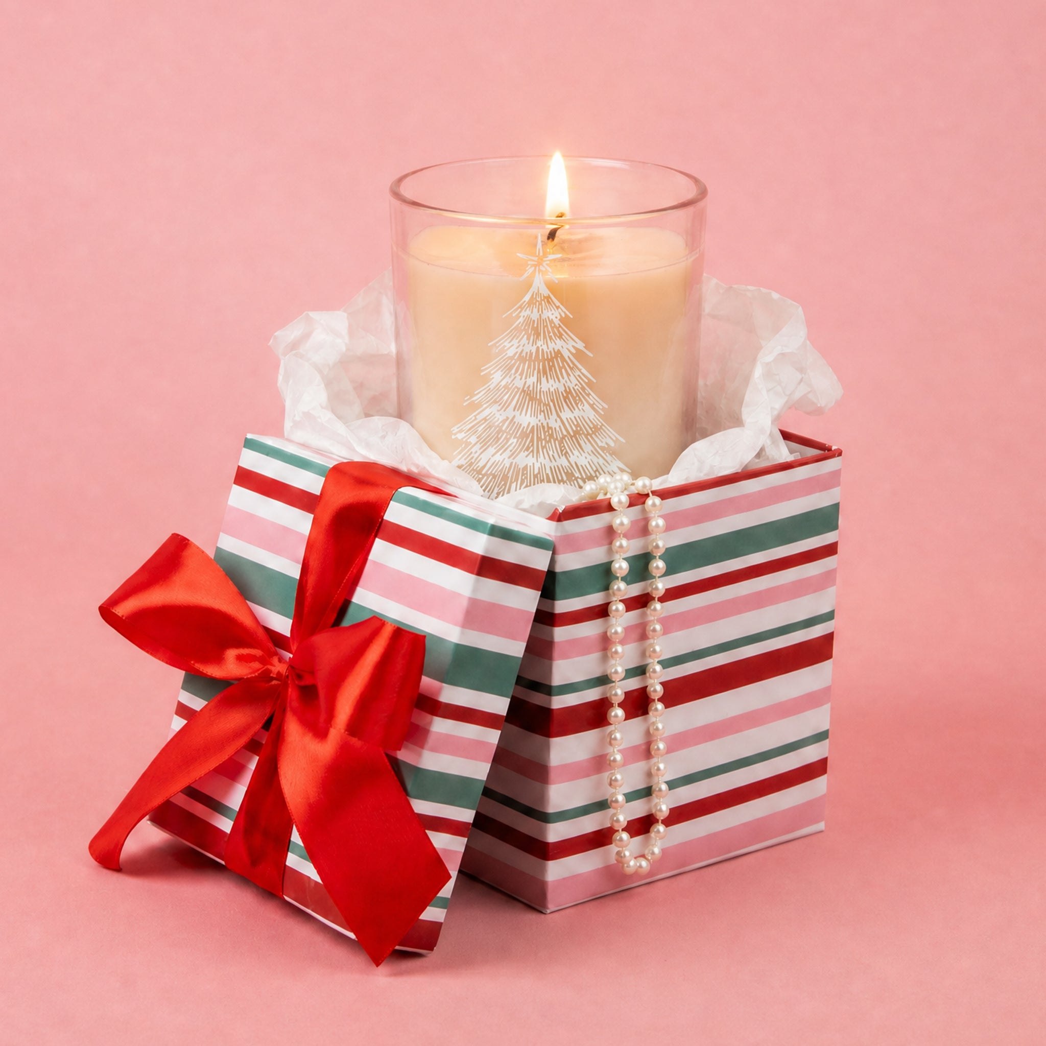A lit Sweet Grace Pink Tree Decal Jar Candle by Bridgewater Candle—with soy-blend wax and a white tree design on the glass—rests inside a striped gift box adorned with a red bow, white tissue paper, and a pearl necklace draped over the edge, all set against a pink background. This festive candle brings elegance and charm to any setting.