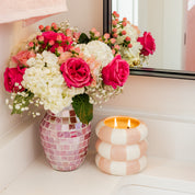 A pink mosaic vase with pink roses, white hydrangeas, and berries sits on a bathroom counter next to a Bridgewater Candle Sweet Grace Collection Candle #074, both reflected in a mirror.