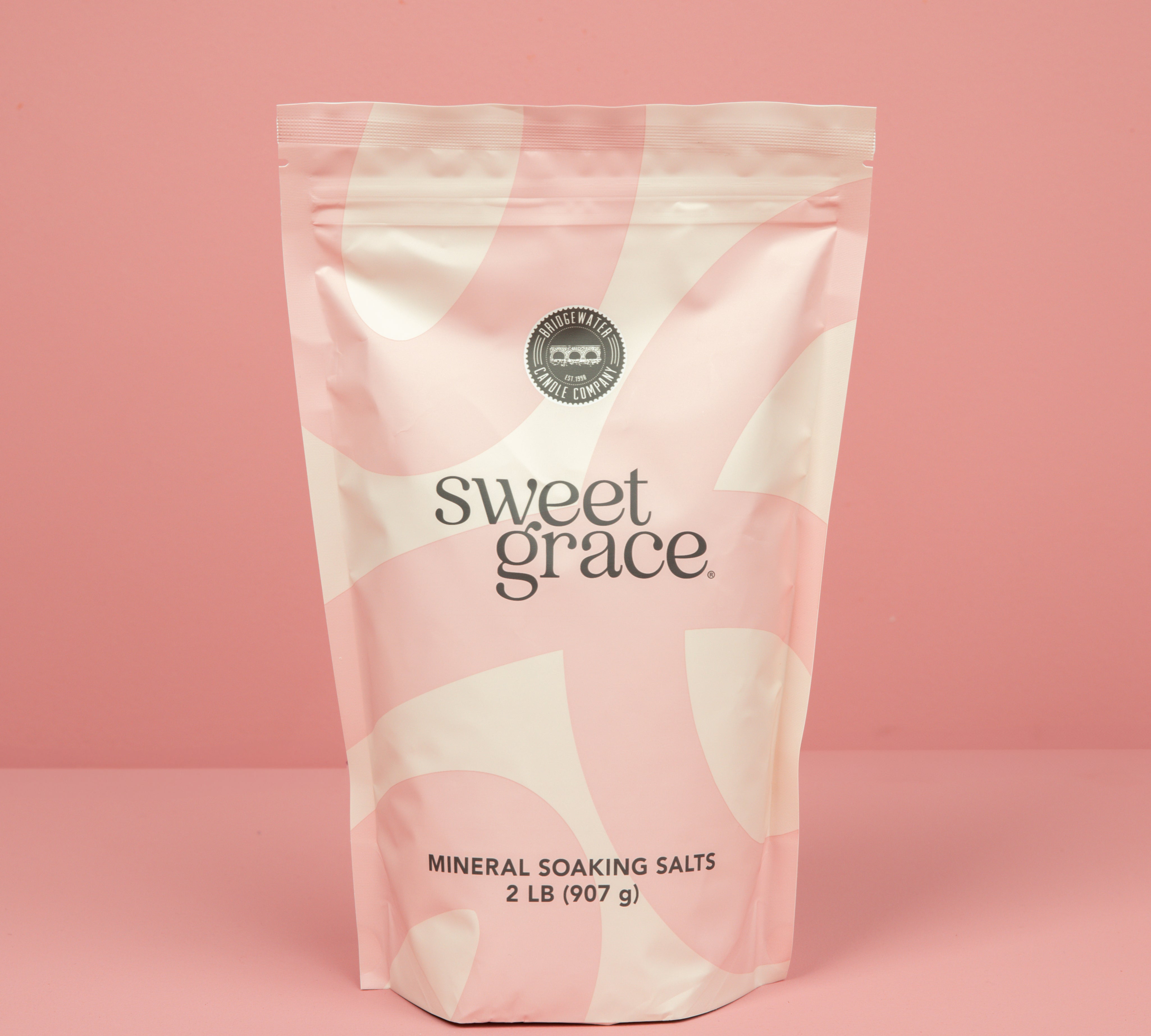 A 2 lb (907 g) pink and white resealable bag of Bridgewater Candle’s Sweet Grace Soaking Salts with Pink Himalayan sea salt stands upright against a solid pink background.