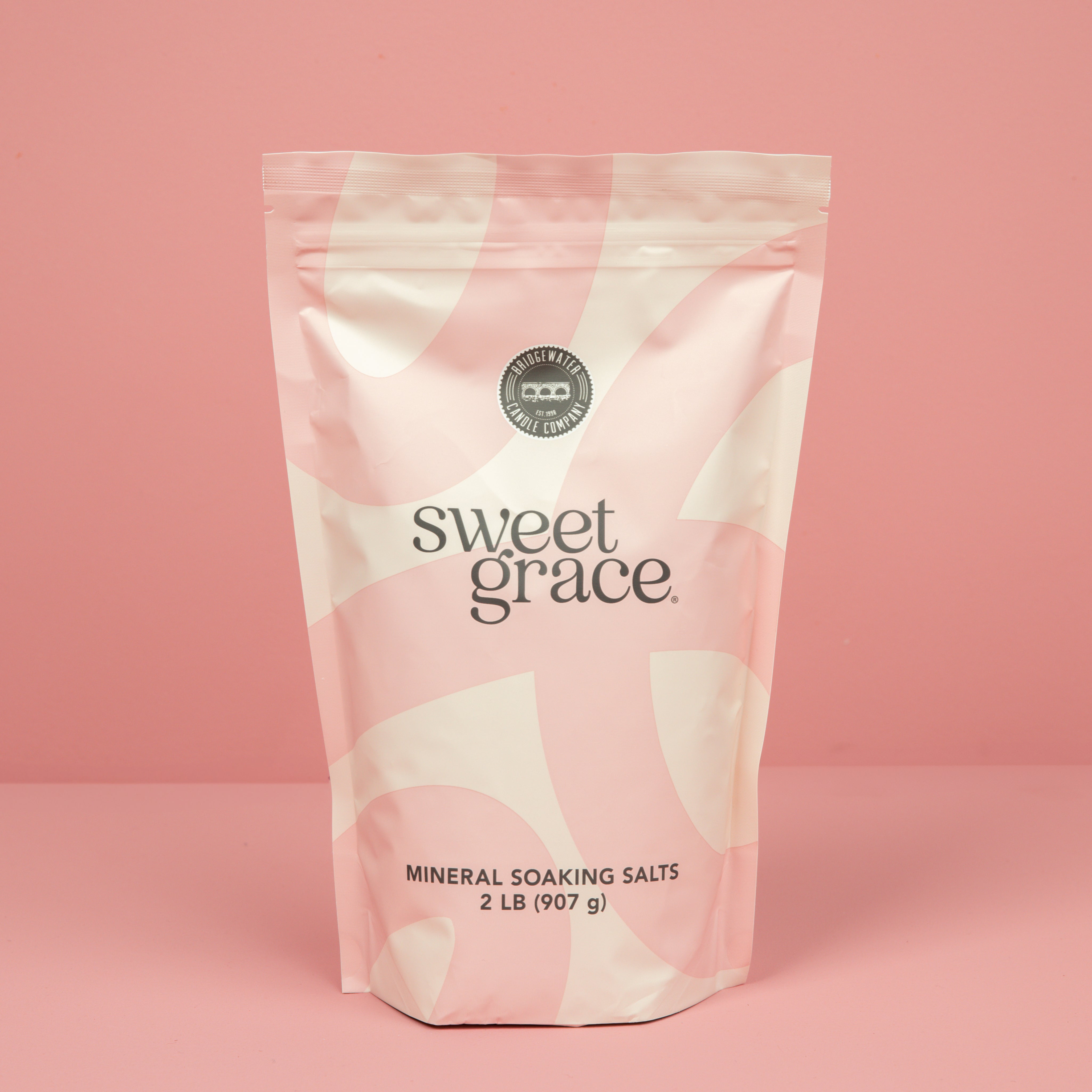 A 2 lb (907 g) pink and white resealable bag of Bridgewater Candle’s Sweet Grace Soaking Salts with Pink Himalayan sea salt stands upright against a solid pink background.