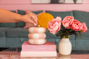 A hand lights the Bridgewater Candle Sweet Grace Collection Candle #074, a striped pink and white soy-blend candle, on a glass table beside pink flowers and a book. A teal sofa with a yellow pillow is in the background.