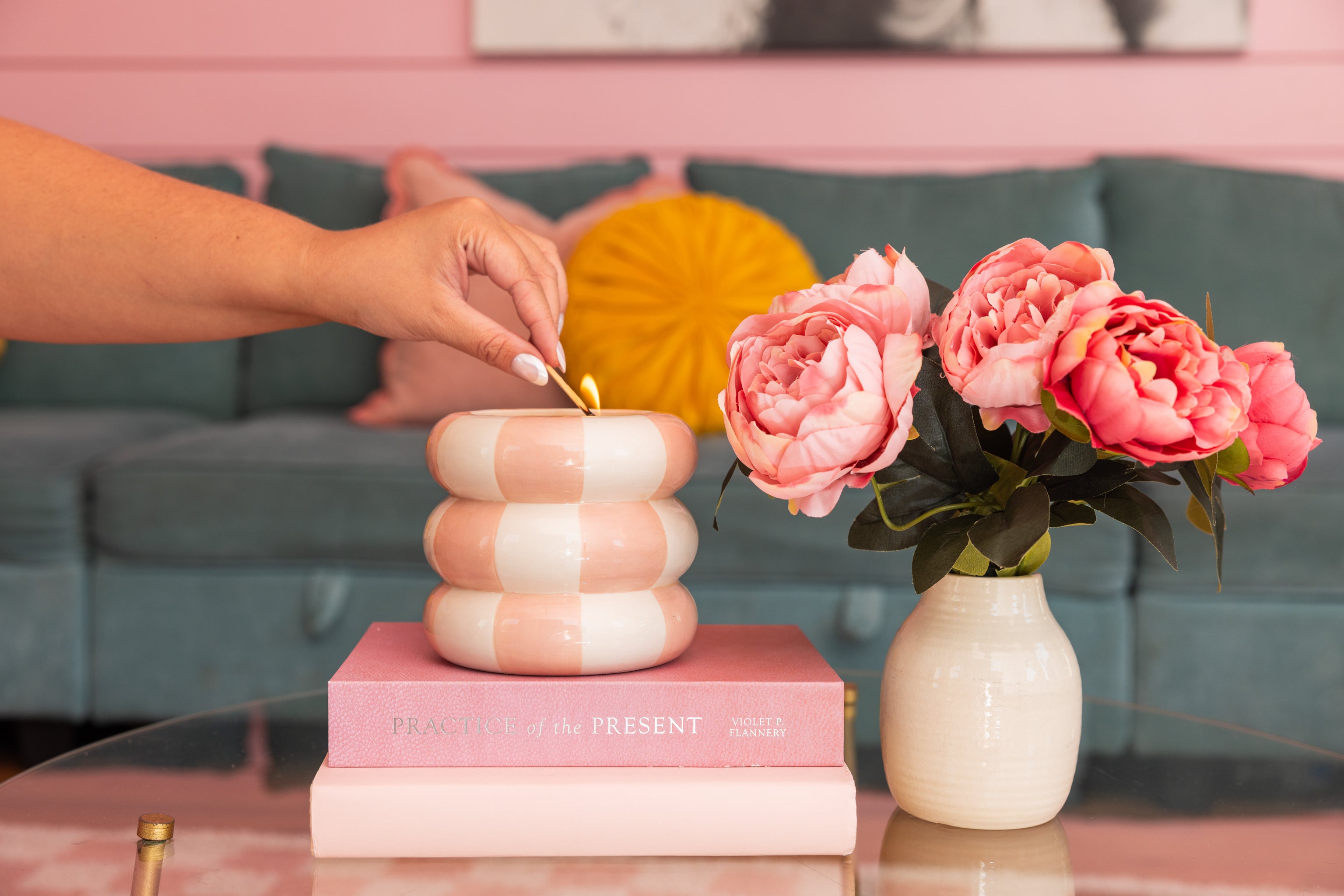 A hand lights the Bridgewater Candle Sweet Grace Collection Candle #074, a striped pink and white soy-blend candle, on a glass table beside pink flowers and a book. A teal sofa with a yellow pillow is in the background.