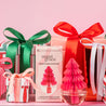 A group of wrapped gifts with red, green, and white ribbons, the Bridgewater Candle Sweet Grace Holiday Honeycomb Diffuser shaped like a pink Christmas tree, and a candy cane are arranged against a pink background—ideal for festive gifting.
