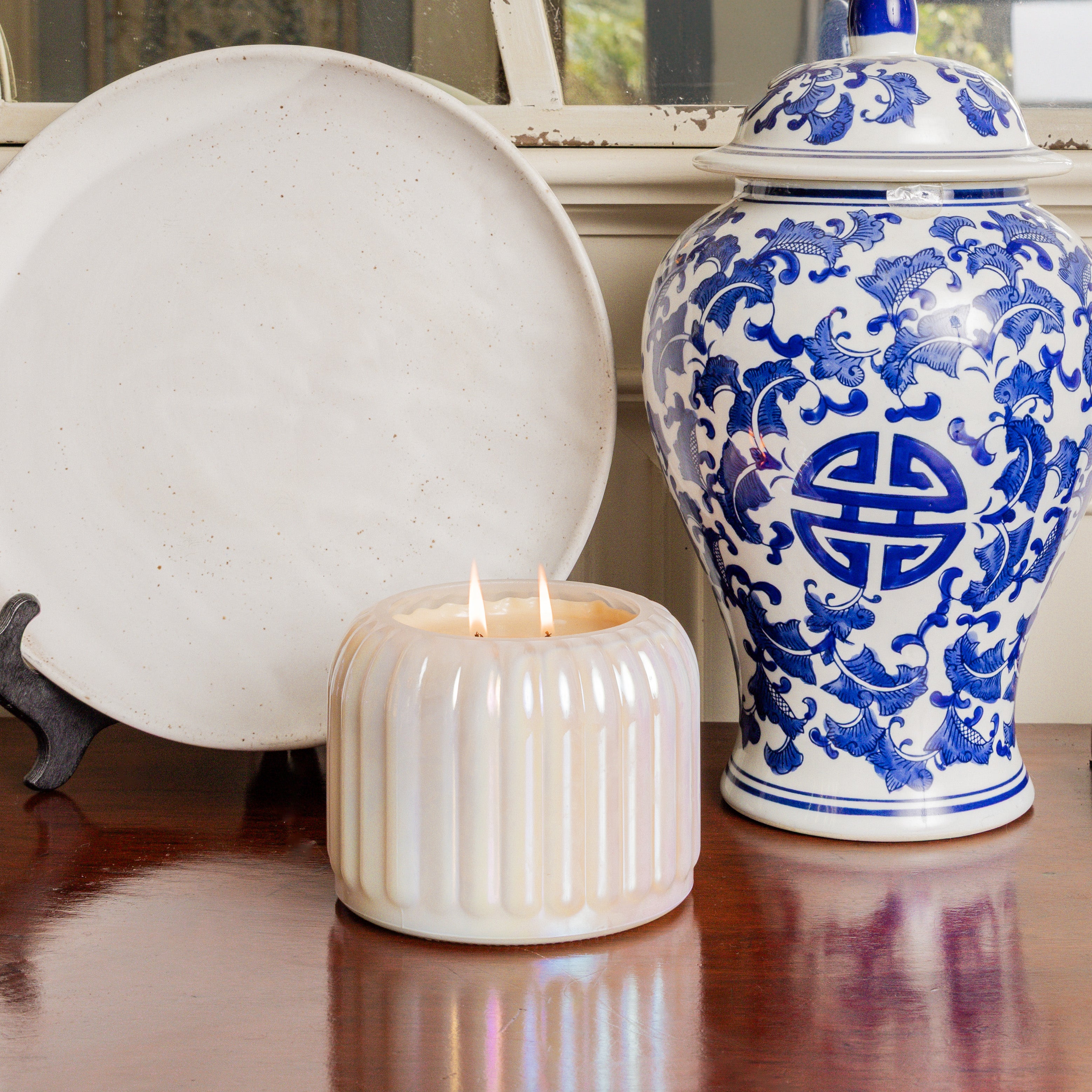 A white decorative plate, a blue and white porcelain jar with patterns, and a lit Bridgewater Candle Sweet Grace Collection Candle #071 with soy-blend wax and lasting fragrance sit on a polished wooden surface.