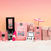 A set of pink Sunkissed fragrance accessories, featuring the Bridgewater Candle Sunkissed 18oz Large Jar Candle, wax melts, and beachy decor like a mini umbrella and surfboard, arranged on sand with a pink gradient background.