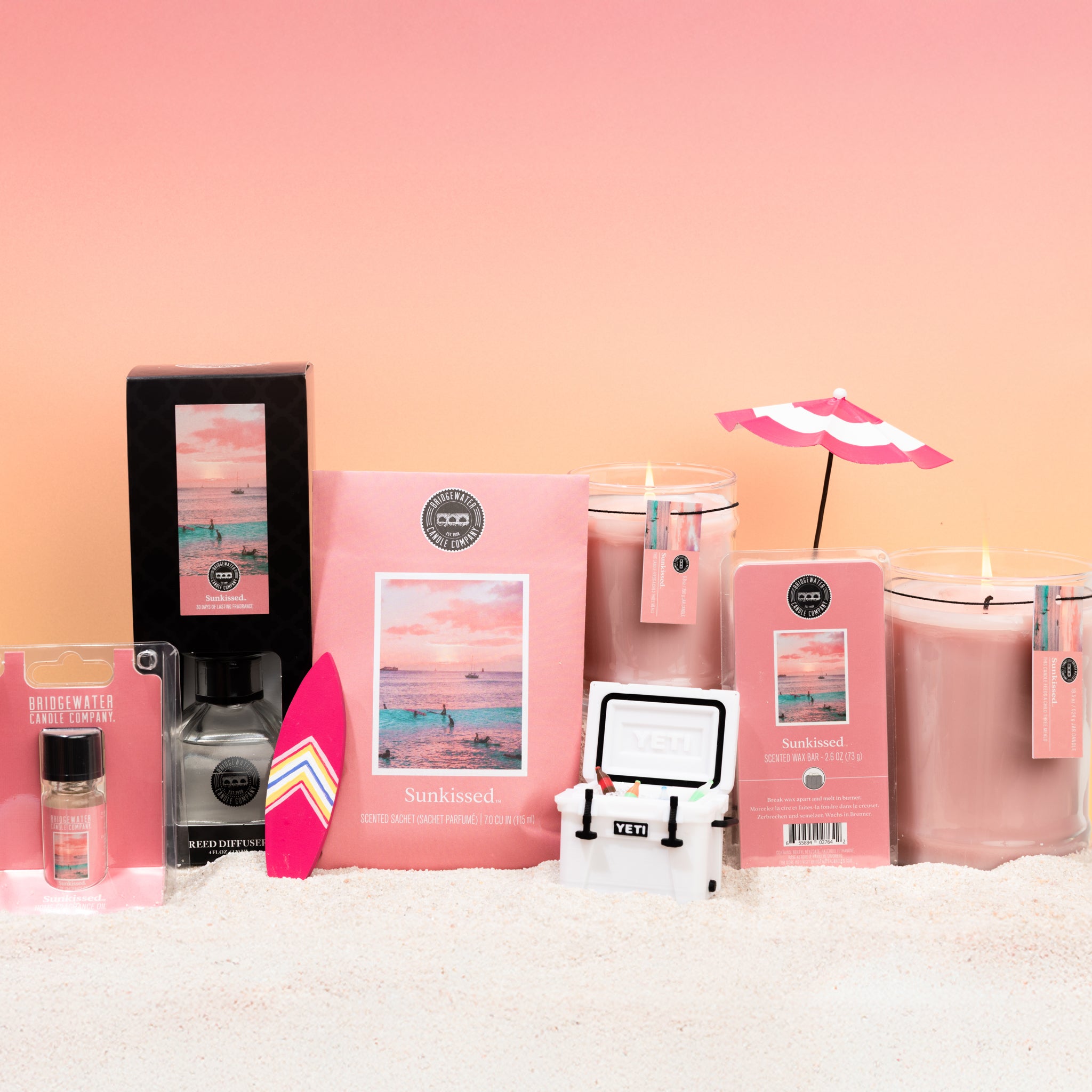 A set of pink Sunkissed fragrance accessories, featuring the Bridgewater Candle Sunkissed 18oz Large Jar Candle, wax melts, and beachy decor like a mini umbrella and surfboard, arranged on sand with a pink gradient background.