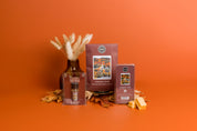 The Bridgewater Candle Autumn Gold Flameless Bundle, featuring a fragrance spray, soy-blend wax bar, and packet, is displayed on an orange surface with dried leaves and a vase of grasses against an orange backdrop.